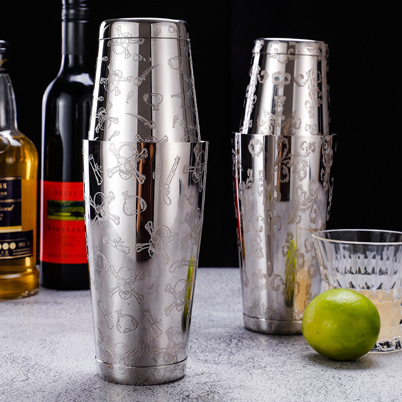 Two metallic cocktail shakers with intricate designs on a bar counter with bottles and a lime.